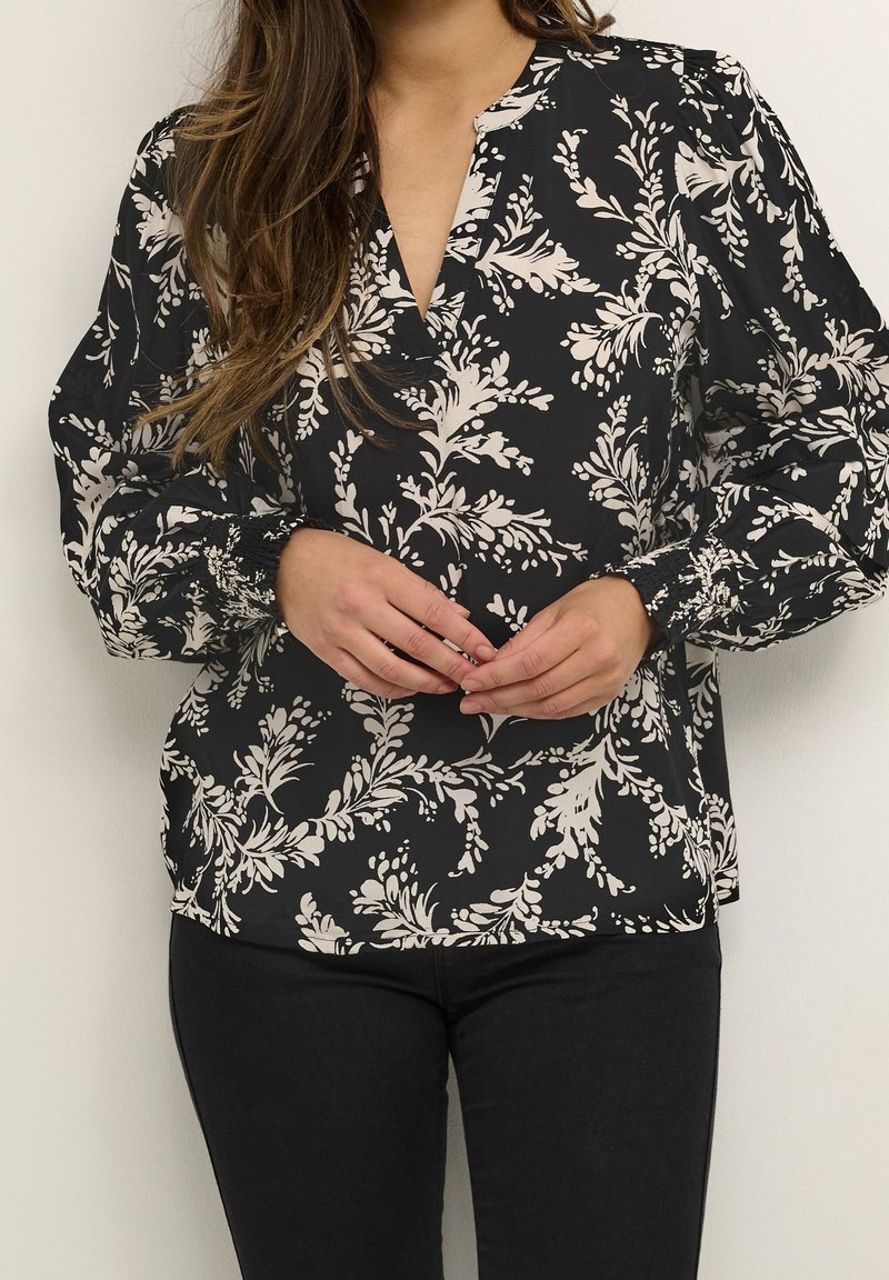 Blouse noire à motif floral blanc, encolure en V et manches longues bouffantes. Le tissu semble léger avec une texture lisse.