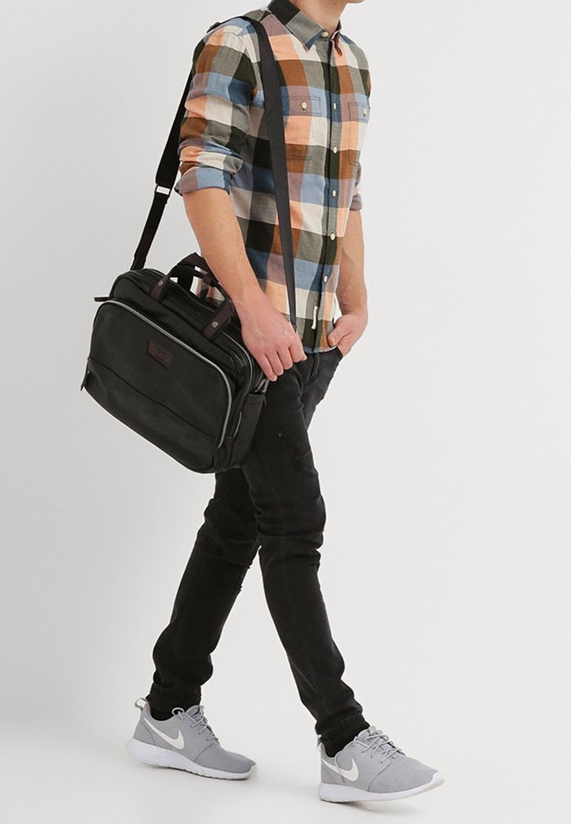 Černá messenger taška s dvojitými ušima a nastavitelným popruhem, nošena s kostkovanou košilí v odstínech modré, oranžové a zelené.