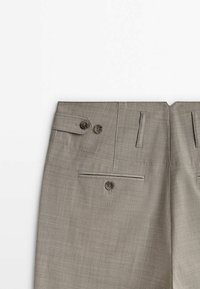 Pantalon habillé marron clair sur mesure, avec poche passepoilée boutonnée à l'arrière et pattes latérales ajustables avec deux boutons.
