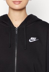 Svart zip hoodie av mjukt material, med frampaneldragkedja, snoddar och en vit Nike-logotyp på vänster bröst.