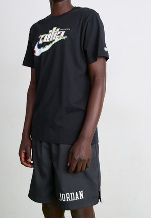 Osoba w czarnej koszulce Nike z kolorowym logo i czarnych szortach Jordana stojąca na jednolitym jasnym tle.