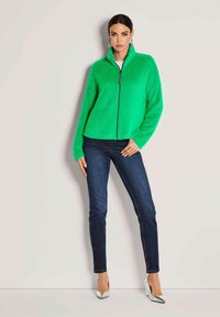 Frau, die an einer Wand steht und eine leuchtend grüne, flauschige Zip-Jacke, dunkle Skinny-Jeans, silberne High Heels und hängende Ohrringe trägt.