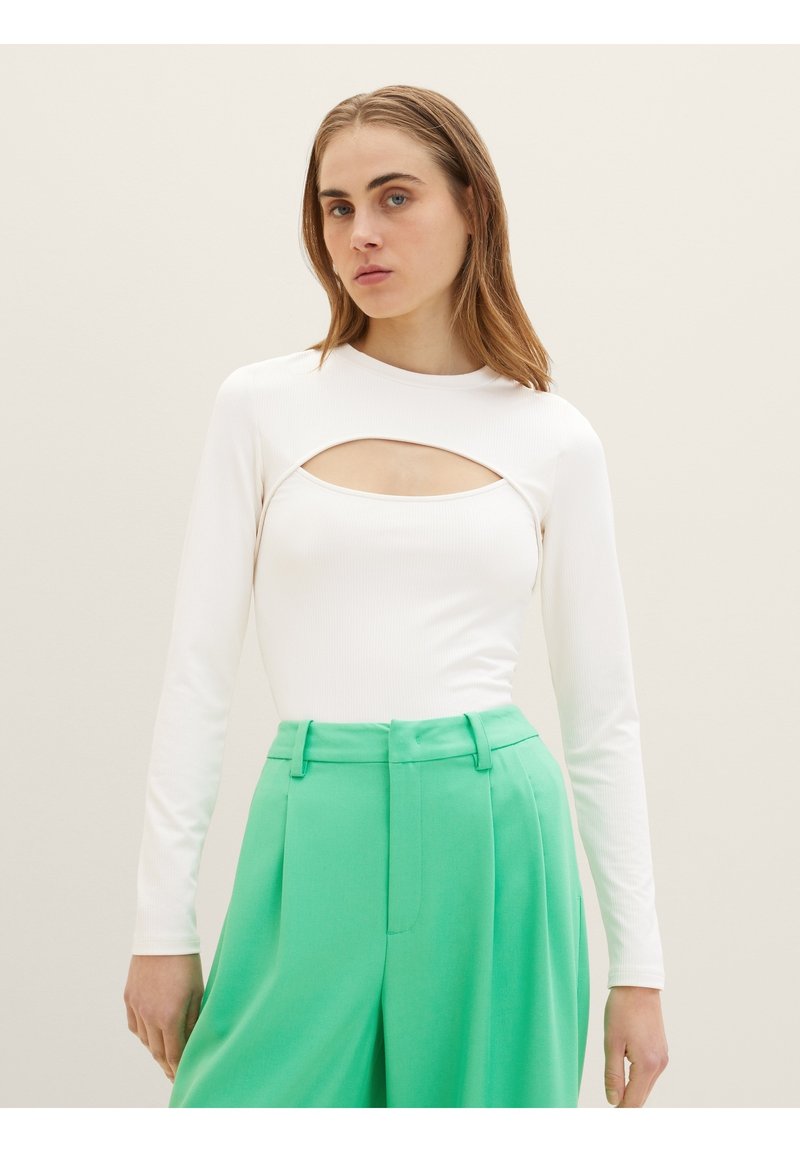 Weißes Langarmoberteil mit einem runden Ausschnitt auf der Brust, kombiniert mit hochgeschnittenen, leuchtend grünen Hosen mit Falten. Glatte Textur.