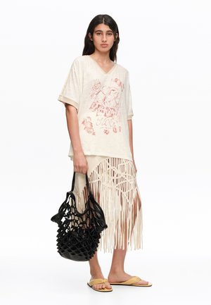 Mujer con camiseta gráfica beige de gran tamaño y falda con flecos, sosteniendo una bolsa negra de red y llevando sandalias amarillas.