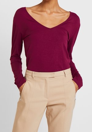 Vrouw die een lange mouwen, bordeauxrode V-hals top draagt, gestopt in beige broek met hoge taille, handen in de zakken tegen een grijze achtergrond.
