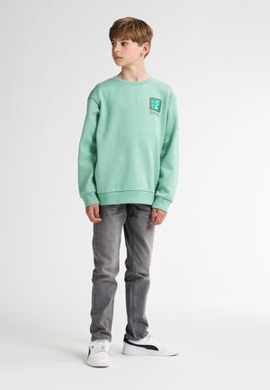 Sweatshirt vert menthe avec des poignets côtelés, présentant une petite étiquette de logo sur la poitrine. Associé à un jean gris et des baskets blanches.