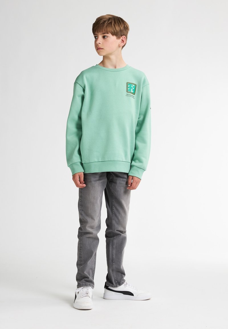Sweatshirt vert menthe avec des poignets côtelés, présentant une petite étiquette de logo sur la poitrine. Associé à un jean gris et des baskets blanches.
