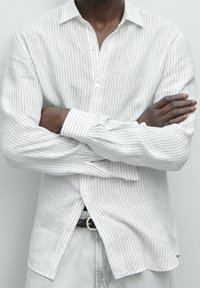 Personne à la peau foncée portant une chemise blanche à fines rayures boutonnée, les bras croisés sur la poitrine, et un pantalon clair avec une ceinture noire.