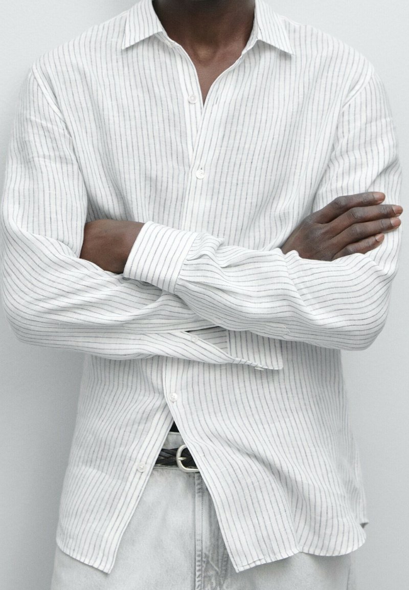 Personne à la peau foncée portant une chemise blanche à fines rayures boutonnée, les bras croisés sur la poitrine, et un pantalon clair avec une ceinture noire.