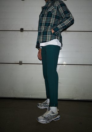 Person trägt türkisfarbene Hose, weiße Socken, silberne Laufschuhe, weißes Shirt unter einem grün-beigen karierten Hemd, steht seitlich.