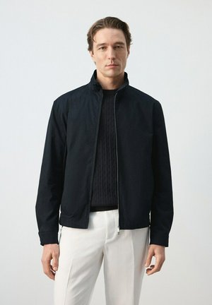 Hombre vestido con una chaqueta negra con cremallera sobre un suéter negro y pantalones blancos, de pie contra un fondo claro y liso.