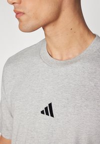T-shirt en coton gris avec un col rond, présentant un logo triangulaire noir brodé sur le côté gauche de la poitrine. Texture lisse.