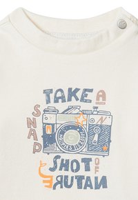 Helles Cremet-Shirt mit einem Grafikmotiv einer Vintage-Kamera und blauen sowie orangen Text, der "Mach ein Schnappschuss von der Natur" lautet.