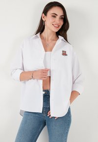 White button-up shirt with a relaxed fit, featuring a teddy bear embroidery on the left chest. Light fabric, long sleeves rolled up.