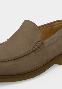 Mocassin en daim marron à enfiler avec couture visible, petit logo embossé sur la tige et semelle en caoutchouc avec doublure intérieure en cuir couleur fauve.