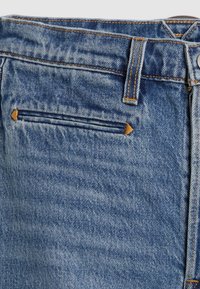 Jean en denim bleu avec une finition texturée, présentant une poche arrière avec une fermeture éclair horizontale ornée d'accents et de coutures en beige.