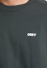 Rövid ujjú, sötétzöld pamut póló kerek nyakkal és egy kis fehér "OBEY" logóval a mellkasán. Sim felület, laza dizájn.