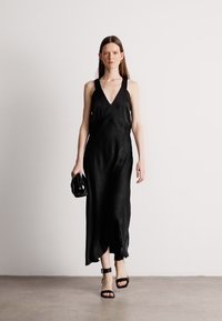 Čierne maxi šaty bez rukávov s hlbokým V-výstrihom, splývavým materiálom a bočným rozparkom. Spárované s malou čiernou kabelkou a čiernymi sandálmi na opätku.