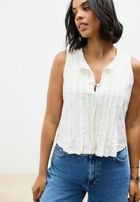Hvid ærmeløs top med vertikal struktur og en nøglehul-udskæring. Sammen med blå højtaljede denimjeans. Enkel design og let materiale.