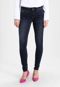 Jeans ajustados de mezclilla en azul oscuro con bolsillos frontales, detalles de costura sutiles y un corte de altura media, combinados con tacones altos rosas.