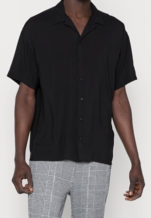 Homme portant une chemise noire à manches courtes boutonnée et un pantalon à carreaux gris, debout devant un fond uni.