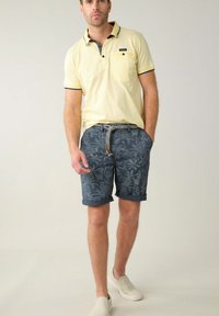 Polo shirt jaune clair avec des accents marine, associé à un short marine avec un motif de feuilles de palmier. Le modèle porte des chaussures slip-on beiges.