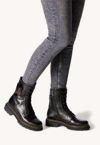 Černé kožené kotníkové boty mají šněrování vpředu, boční zip, přízdobu s přezkou a robustní gumové podrážky, v kombinaci se šedými džíny skinny.