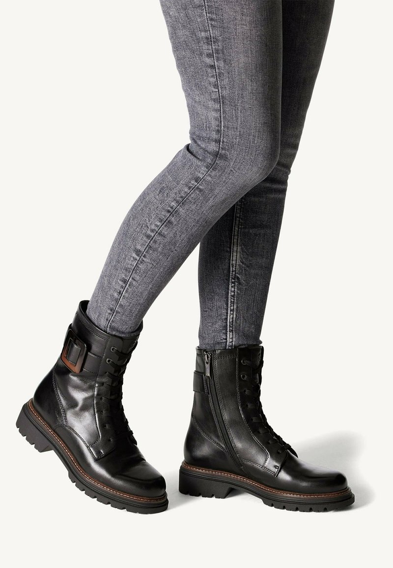 Černé kožené kotníkové boty mají šněrování vpředu, boční zip, přízdobu s přezkou a robustní gumové podrážky, v kombinaci se šedými džíny skinny.