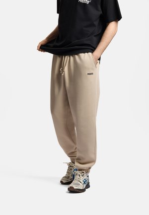 Osoba nosząca beżowe spodnie dresowe z sznurkiem, czarną koszulkę i białe trampki z niebieskimi i czarnymi detalami, stojąca na białym tle.