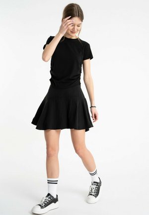 Młoda kobieta poprawia włosy, ubrana w czarną koszulkę, czarną spódnicę, białe skarpetki w paski i czarno-białe trampki na białym tle.