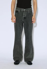 Osoba ubrana w czarne, wysokie, szerokie dżinsy w odcieniu wyblakłym, czarny top na ramiączkach i czarne sneakersy z białymi przeszyciami. Ręce w luźnej pozycji po bokach.