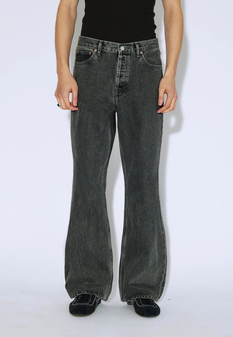 Osoba ubrana w czarne, wysokie, szerokie dżinsy w odcieniu wyblakłym, czarny top na ramiączkach i czarne sneakersy z białymi przeszyciami. Ręce w luźnej pozycji po bokach.