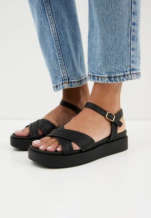 Voeten met zwarte platformsandalen met enkelbandjes en lichtblauwe spijkerbroek die boven de enkels is afgeknipt, op een witte vloer.