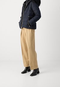 Doudoune marine avec capuche bordée de fourrure, associée à un pantalon plissé beige et des bottines à talons carrés noires, ornées de ferrures argentées.