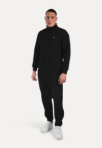 Joven de pie vistiendo un sudadera negra de medio cierre, pantalones deportivos negros y zapatillas blancas frente a un fondo claro y liso.