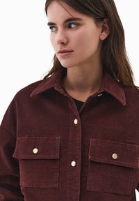 Bordowa koszula z korduroju z dużym kołnierzem, dwiema przednimi kieszeniami i złotymi guzikami. Miękka tekstura i luźny krój.