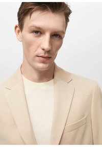 Blazer beige con tessuto testurizzato, taschino sul petto, indossato sopra un maglione crema chiaro. Colori neutri, vestibilità su misura, design moderno.