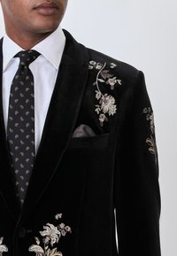 Man wearing black velvet blazer with white floral embroidery, white dress shirt, and black paisley-patterned tie.