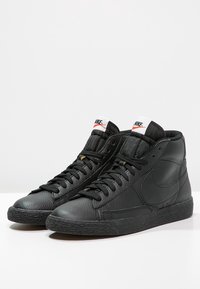 Schwarze Leder-Hochschnitt-Sneaker mit strukturierten Oberflächen, schwarz Schnürsenkel, einem gepolsterten Kragen und einem weißen sowie orangefarbenen Nike-Logo auf der Zunge.