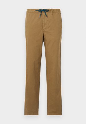 Mies, joka pukeutunut valkoiseen Paul Smith -t-paitaan, jossa keltainen logo, ruskeat kiristettävät housut, mustat kengät, pitelee mustaa käsilaukkua sivullaan.