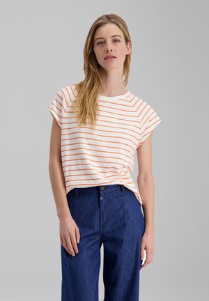 Blonde Frau trägt ein weißes T-Shirt mit orangenen Streifen und hoch taillierte blaue Jeans, steht vor einem grauen Hintergrund.