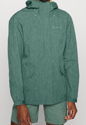 Grøn hættejakke til udendørs brug med forlommer båret af en person i matchende grønne shorts mod en ensfarvet baggrund.