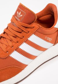 Orange sneaker med mocka- och textilmaterial, tre vita ränder och en strukturerad vit sula. Inkluderar varumärke på plösen.