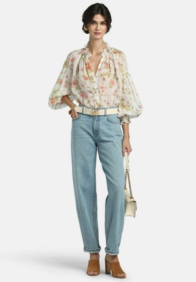 Kvinde iført en gennemsigtig blomstret bluse med puffede ærmer, højtsiddende lyseblå jeans, beige hæle, hvidt bælte og bærende på en lille beige håndtaske.