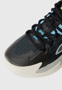 Svart och blå sneakerspets med zigzag-mönster, svarta snören, blå logodetaljer och vit texturerad sula mot en enkel bakgrund.