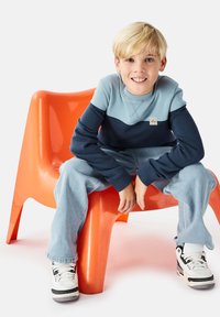 Sininen ja tummansininen värilohkoilla varustettu collegepaita, jossa on resorit hihansuissa, yhdistetty vaaleansinisiin farkkuihin. Istuu kirkkaan oranssilla muovituolilla.