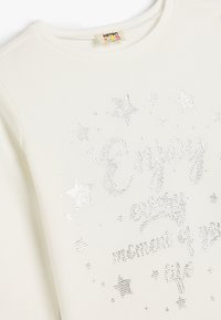 Hvid langærmet bluse med ribbet tekstur. Har metallic sølvtekst og stjernedesign, som formidler et legende og muntert budskab.