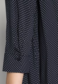 Navy blue polka dot shirt with white dots, featuring a buttoned cuff and soft fabric texture, capturing a close-up of the sleeve design.