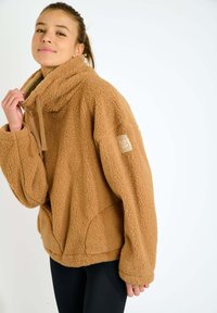 Pullover in pile marrone, con colletto alto, grande tasca anteriore e etichetta sulla manica. Materiale morbido e testurizzato con vestibilità ampia.