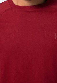 T-shirt rossa in cotone con scollo tondo, cuciture visibili e un discreto dettaglio del logo sul petto. Texture liscia, design casual.